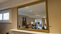 Full view of large rectangular mirror hung on wall reflecting part of room with window shutters and artwork.
