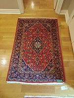 Full view of red machine-made Persian-style rug on hardwood floor showing overall dimensions, central medallion and white fringe tassels.