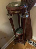 Full view of wood plant stand showing large round green marble top, brass floral trim, and winged lion head details on tripod legs.