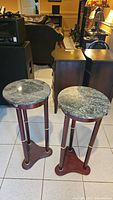 Two round Bombay stone top plant stand tables with wooden legs and brass accents shown from above and front, placed on tiled floor in a living room setting.