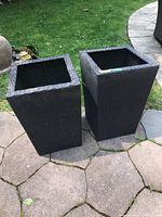 Two tall square black faux wicker planters placed outdoors on stone pavement and grass, showing their exterior and overall shape.