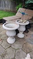 Two resin urn planters on stone patio, one containing metal bird and dragonfly garden decor.