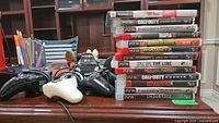Controllers and game cases displayed on a table, showing two PS2 controllers (black and white), one Chillstream PS3 controller, one PS2 game, and a stack of 12 PS3 game cases.