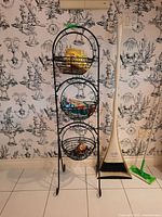 Front view of black metal basket rack with three circular baskets filled with food packages, placed against wall with black and white wallpaper. Adjacent to white broom and green mop.