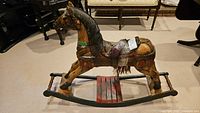 Side view of wooden rocking horse showing detailed paintwork and attached saddle, with some damage visible.