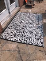 Full view of rectangular outdoor rug with black and white diamond geometric pattern placed on stone floor outdoors near glass doors.