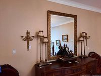 Pair of decorative metal sconces mounted on a wall with a large gold framed mirror and vintage lamps on a wooden dresser beneath.