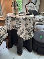 Square plywood table draped with black and white toile-patterned fabric, showing part of upholstered furniture around.