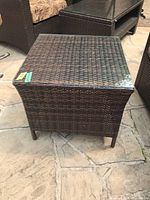 Front view of the faux wicker side table with glass top outdoors on a stone patio next to outdoor furniture.
