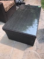 Faux wicker table with glass top shown on outdoor stone patio from a top angle. Reflections visible on glass surface.