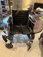 Front view of the black manual wheelchair showing padded seat and armrests, small front wheels, and one footrest removed on tiled floor.