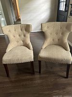Front view of two beige fabric tufted contemporary accent chairs with wooden legs and silver nailhead trim, showing button tufting on backrest and curved wingback side design.