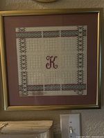 Framed cross stitch artwork showing the letter 'K' with colored border, mounted and framed.