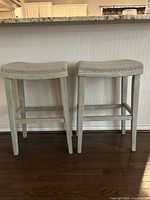 Two gray wooden counter stools side by side in front of a white paneled kitchen counter with granite top, showing the full height and rectangular seats with studded trim.