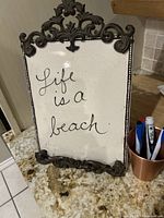 Front view of the 'Life is a beach' decorative sign showing the ceramic panel with cursive black writing and ornate metal frame.