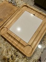 Large wooden cutting board with natural wood finish, embedded white ceramic insert, two circular recesses, and edge groove, photographed on granite counter.