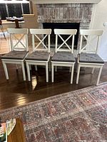 Front view of four white wooden dining chairs with X-back design lined up in a row on a dark hardwood floor in front of a fireplace. Each chair has a gray button-tufted cushion on the seat.