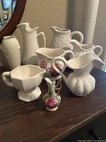 Seven white ceramic pitchers of various traditional designs displayed on a wooden surface showing shapes, handles, and decorative details including a small floral patterned piece.