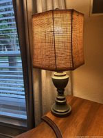 Photo of the complete table lamp illuminated, showing the burlap fabric shade and wooden base with decorative curves and a distressed blue-gray finish.