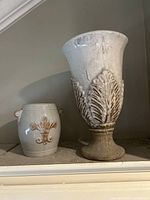 Two cream-colored vintage pottery pieces with fleur-de-lis and leaf patterns, one pot with handles and one large footed vase.