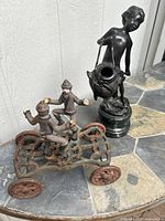 Photo showing two cast iron figurines: a dark patinated boy statue with a basket, and a rusty multicolor wheeled toy with two characters.