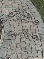 Two ornamental wrought iron decorative panels displayed on a paved walkway showing intricate scroll designs in medium and large sizes.