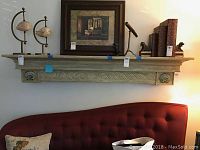 Full view of wooden mantel shelf mounted on wall above red tufted couch. Shelf decorated with framed print, shell sculptures, small telescope, bookends with dog figurines, and books.
