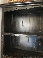 Close-up view of the upper open shelves of the black distressed cupboard showing scalloped trim and wear marks on the wood surfaces.