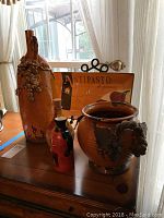 Photo of a globular decorative wine vessel with grape clusters, Antipasto plaque with metal stand, and two decorative bottles on a wooden table near a window.