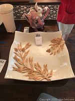 Photo showing large square cream-colored platter with raised berry and leaf design on the edge, placed on wooden table with other decorative items visible.