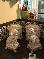Pair of lidded oval porcelain jars with matching floral and polka dot patterns and green handle lids, accompanied by two matching seated dog figurines with pink collars, sitting on bases with green and gold accents on a dark countertop with mosaic tile backsplash.
