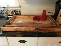 Two nested wooden trays with natural finish and side handles on kitchen counter.