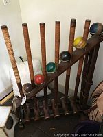 Full view of six wooden croquet mallets with wrapped handles and six differently colored balls arranged on a wooden rack.