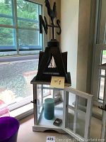 Decorative candle house with glass door open, blue pillar candle and small lighter inside, natural light from window behind