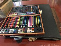 Top view of open wooden art set showing compartments filled with colored pencils, crayons, watercolor paint trays, brushes, charcoal sticks, and erasers. Partially visible black portfolio and black desk pad beneath the art set.