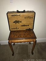 Front view of a vintage suitcase with painted fish designs and ornate wooden tea table below it.