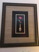 Front view of framed floral wall art showing red flower in vase on black board and large giltwood frame.