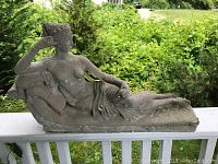 Grey stone-like reclining female statue shown from side view, placed on white railing with green bushes in background