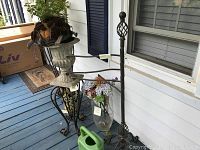 Wrought iron plant holder, stone urn planter, glass vase with flowers, and green watering can on blue wooden porch floor by window with dark blue shutters.