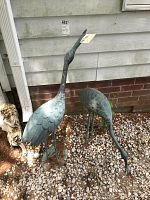 Two cast metal water bird sculptures outdoors on gravel, one taller with neck raised and one smaller with neck down.