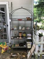 Full view of the metal outdoor bakers rack with tile shelves outside near a porch area, showing its overall design and condition.