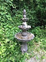 Full height view of the tall resin fountain showing two scalloped basins and the ornate but chipped top finial, surrounded by greenery and gravel ground.