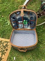 Open wicker picnic basket showing blue and white gingham fabric interior lining and picnic set components secured in lid, including blue cups, plates, and cutlery wrapped in gingham cloth.
