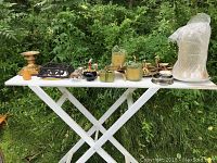 Photo of white table outdoors showing various candle holders, candles, decorative and lighting accessory items arranged on a foldable table with greenery background.