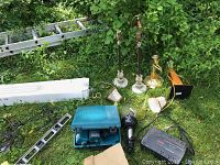 Photo showing Makita drill in case, several lamps with shades, APC battery backup, metal level, decorative metal wall hangings, and ladder in grass.