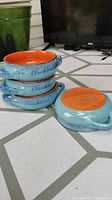 Four stacked ceramic ramekin bowls showing blue exterior with brushstroke pattern and bright orange interior, along with one bowl laying on side to show underside and handle.