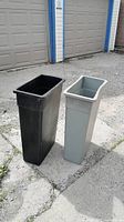 Two rectangular garbage bins, one black and one gray, standing outside on a concrete surface against a backdrop of garage doors. Both bins are empty and upright, showing narrow rectangular shapes with open tops.