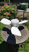 Photo of a white Honeywell ceiling fan with four blades and integrated light fixture set on a round wooden outdoor table, with grass and garden background.