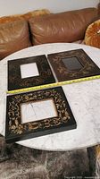 Three black wooden picture frames with hand painted gold floral decorations on a white table with a brown couch in background