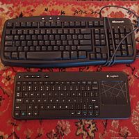 Photo showing both keyboards placed on a carpet. The Microsoft wired keyboard features a standard full layout, while the Logitech wireless keyboard has a touchpad on the right side and additional media keys.